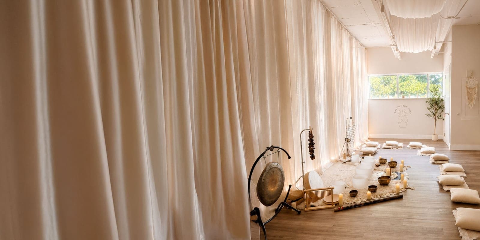 The Balance You Seek studio set for a sound bath: a long wall of sheer ivory curtains lit warm from behind, a gong and chimes beside crystal singing bowls and candles on sheepskin, and floor cushions lining the wood floor toward a bright window and olive tree.